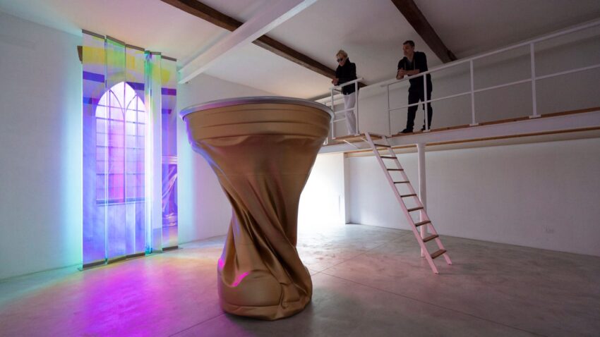 Two people stand on a mezzanine looking at a large, gold, twisting sculpture in a modern art gallery with colorful lighting.