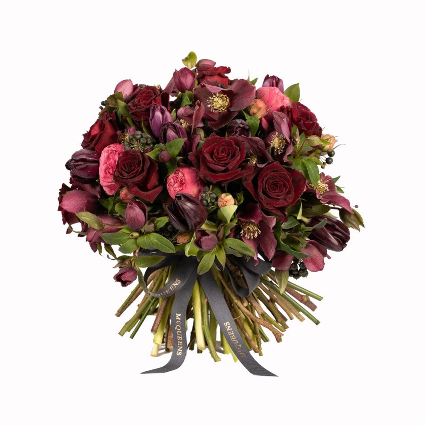A bouquet of red roses and dark purple flowers with greenery, wrapped with a black ribbon, arranged in a round shape.