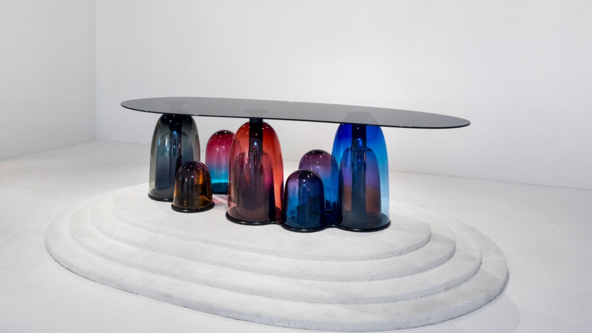 Modern glass table with colorful, abstract glass base on a white tiered platform in a minimalist room.