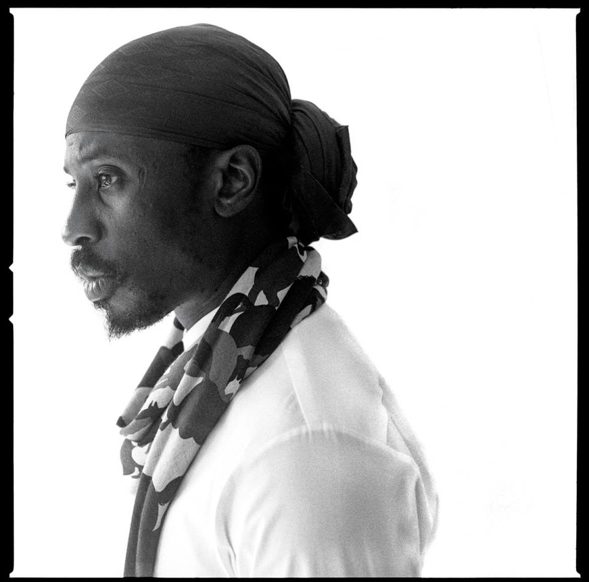 Man wearing a headscarf and camo scarf, turned to the left against a plain background, displaying a thoughtful expression.