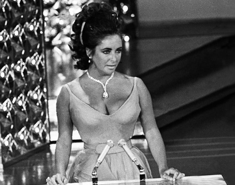 Woman in an elegant dress speaking at a podium with microphones, wearing a diamond necklace, black and white image.