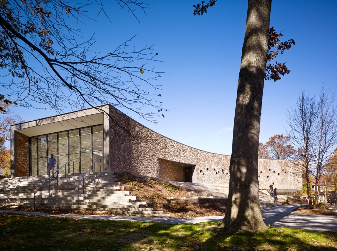 Modern building with large glass windows and brick exterior, surrounded by trees and a clear blue sky in a park setting.