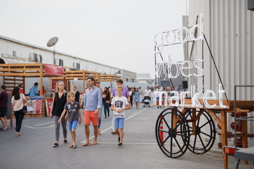 People walking at an outdoor market near a neon sign that reads "good vibes market" with stalls and a casual atmosphere.