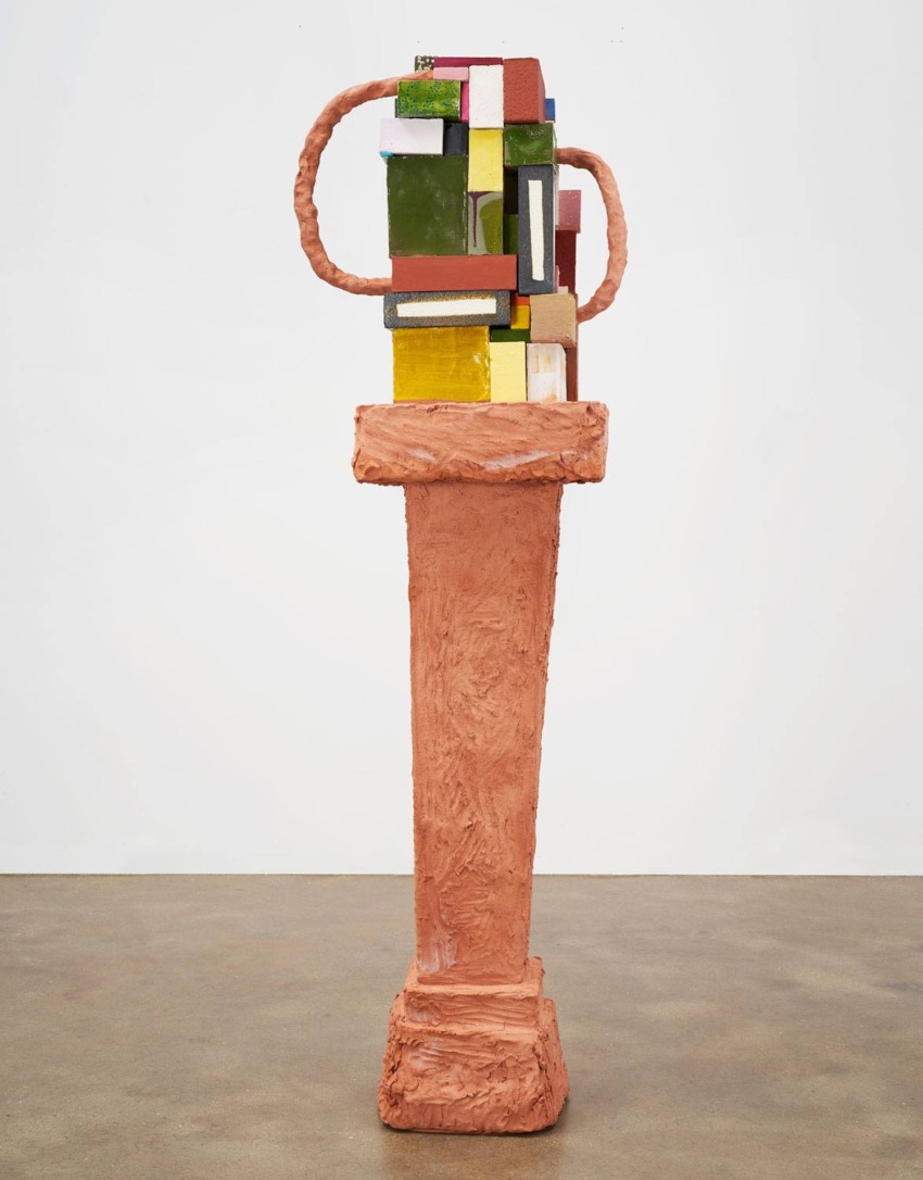 Colorful abstract sculpture on a tall textured pedestal, featuring geometric shapes and earthy tones against a plain background.