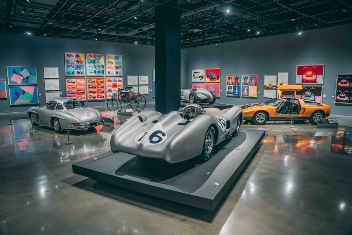 Classic car exhibition with vintage silver race cars and an orange car displayed in a modern showroom setting.