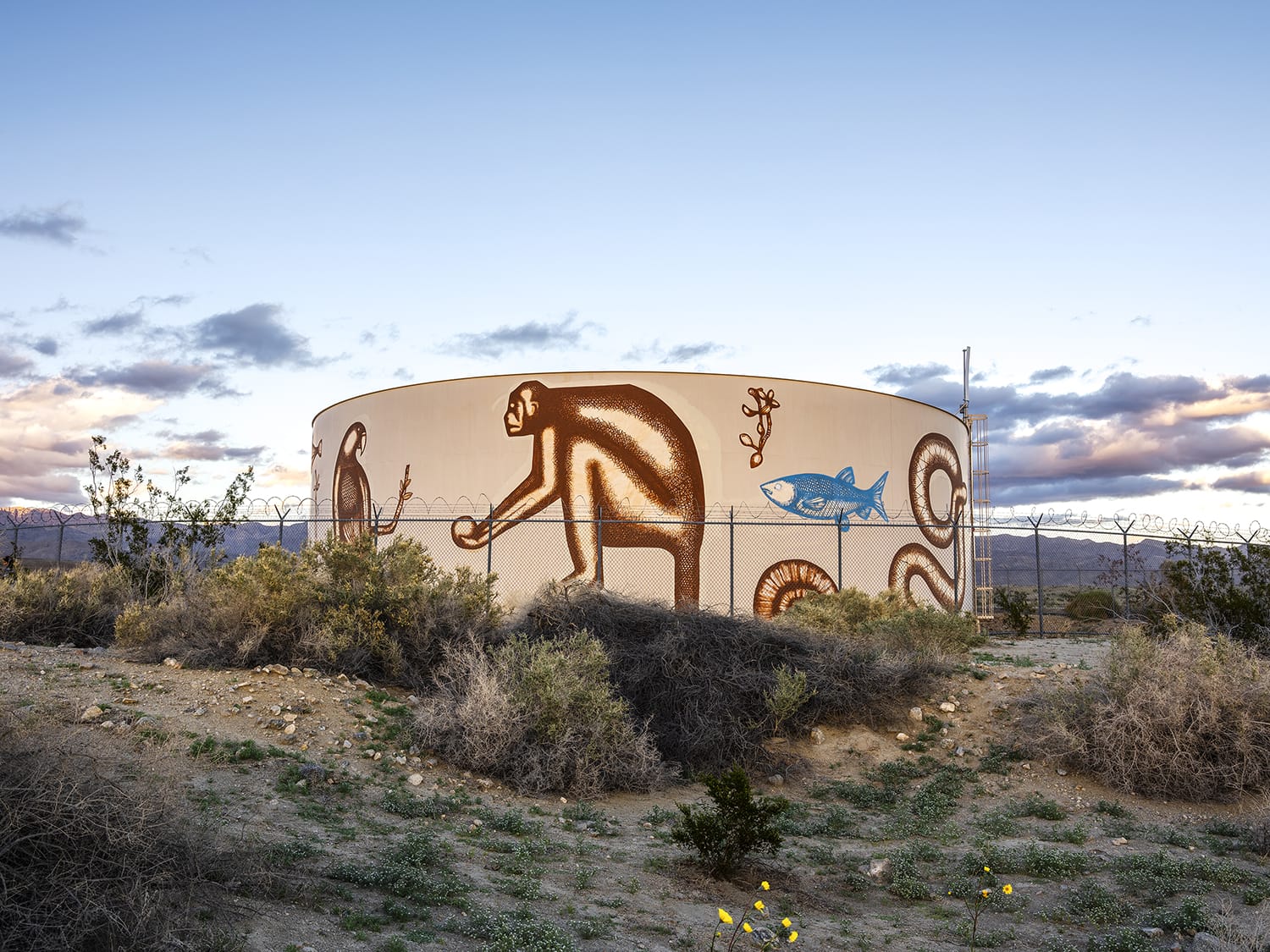Water tank with colorful animal murals in a desert landscape under a cloudy sky.