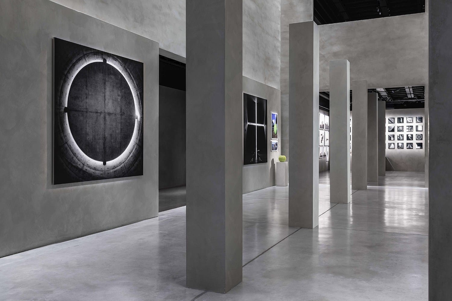 Contemporary art gallery interior with concrete walls, high ceilings, and abstract paintings displayed on pillars and walls.