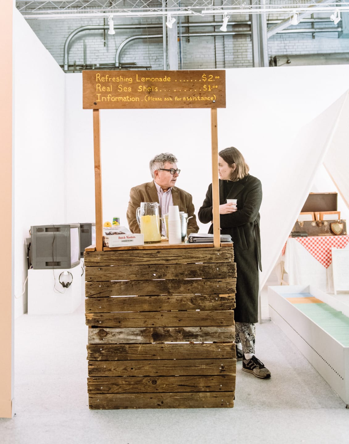 Man and woman at a rustic lemonade stand with a sign listing prices for lemonade and sea shells in an indoor setting.