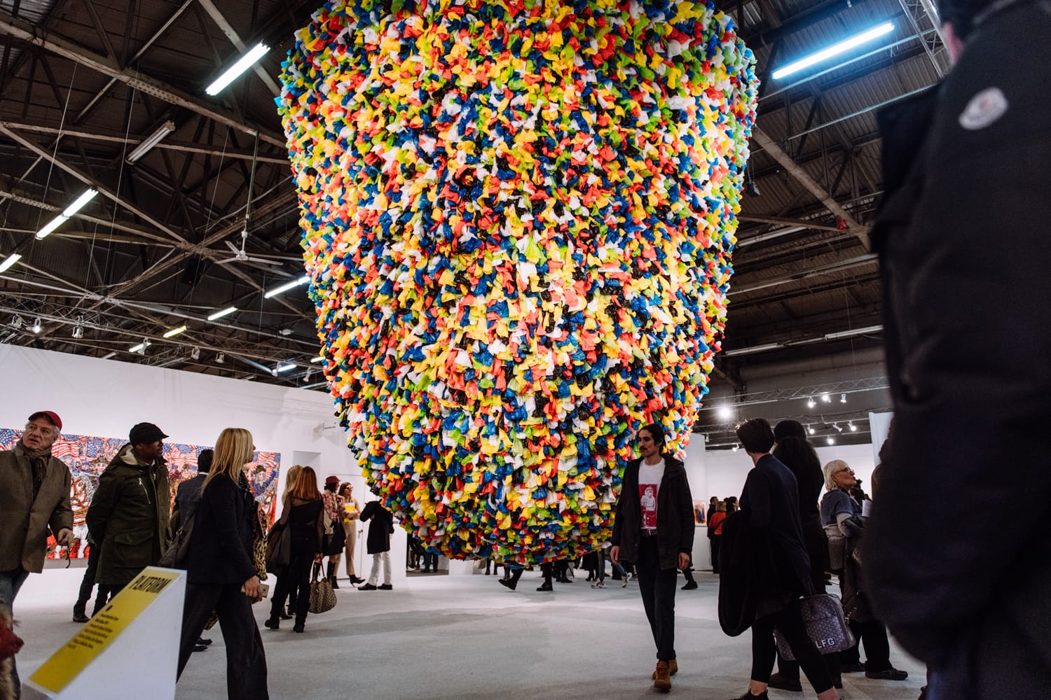 Visitors admire a large, vibrant art installation made of colorful fabric pieces suspended from the ceiling of a gallery.