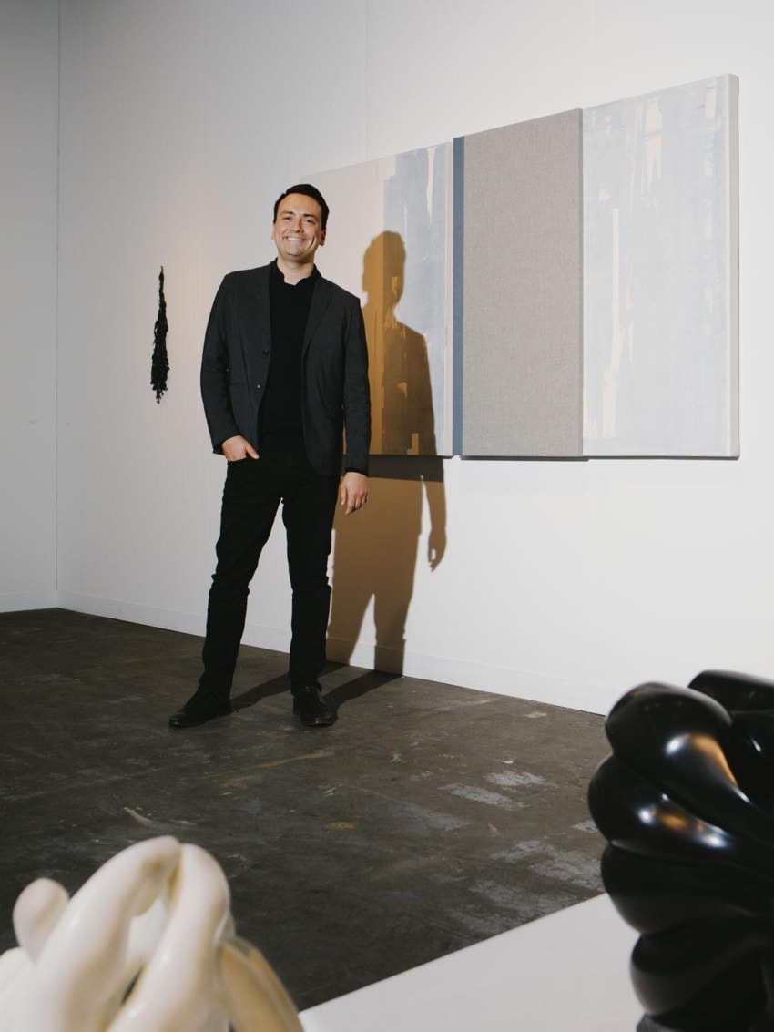 Person standing in a gallery, smiling, with modern art on white walls and sculptures on display.