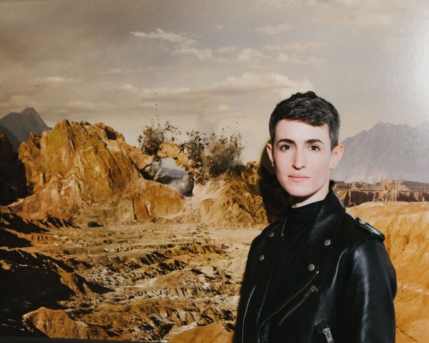 Person in a leather jacket stands in front of a rocky landscape with mountains and an explosion in the background.