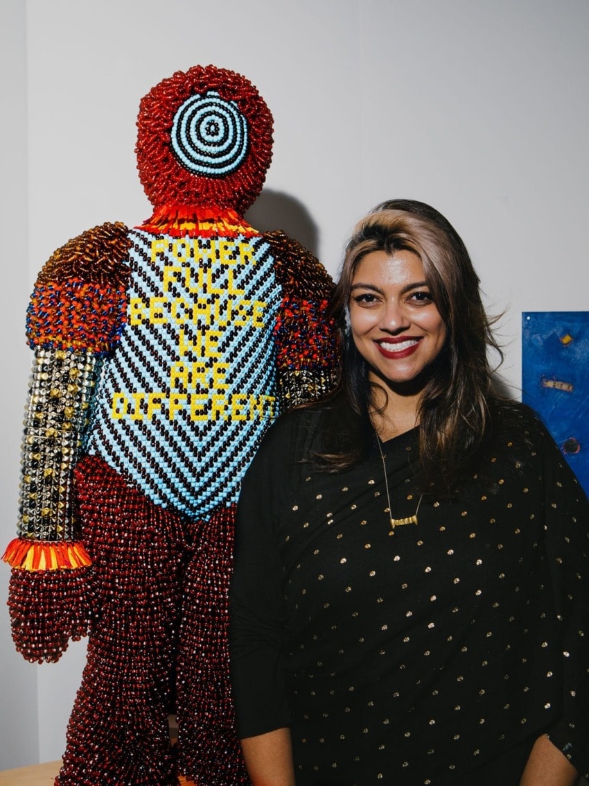 Person standing next to a colorful beaded sculpture, smiling at the camera, with artwork in the background.