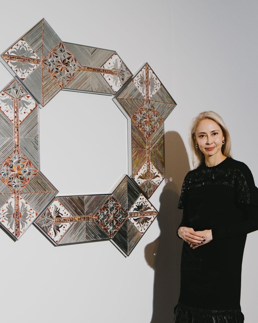 Woman standing next to a geometric art piece on a white wall, featuring intricate patterns and metallic elements.