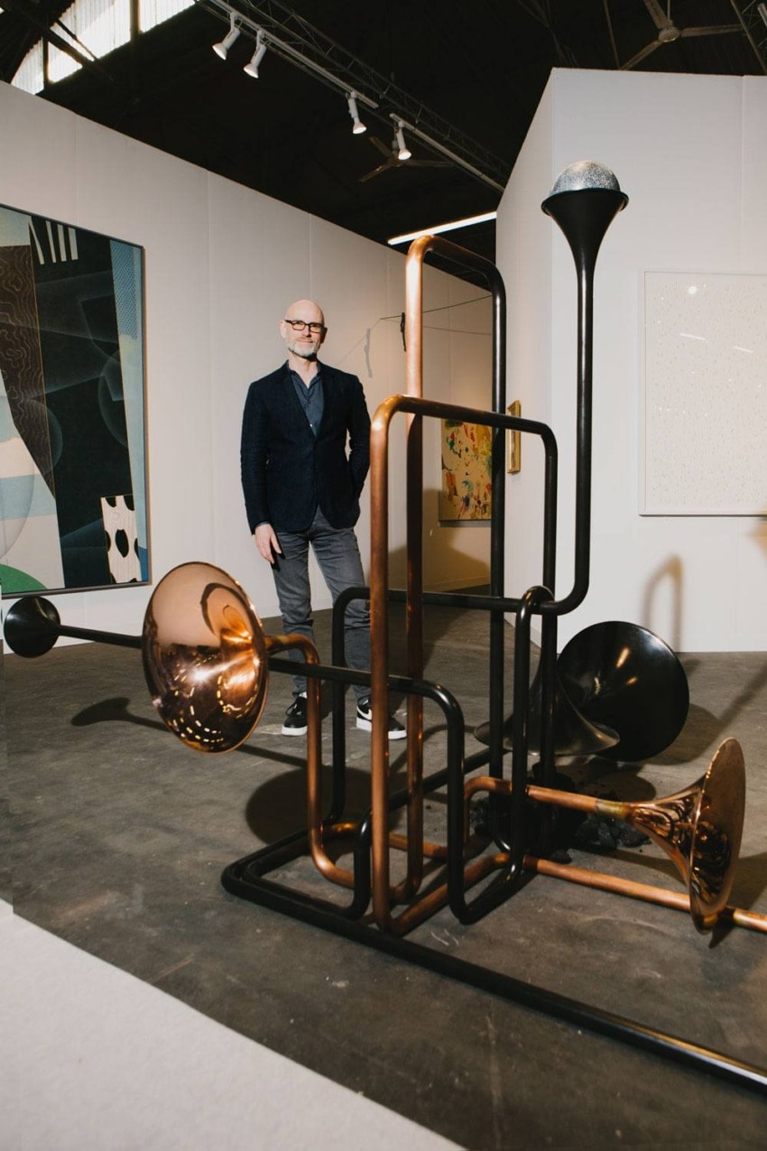 Man standing in an art gallery beside a large abstract sculpture with brass elements and various artworks on the walls.