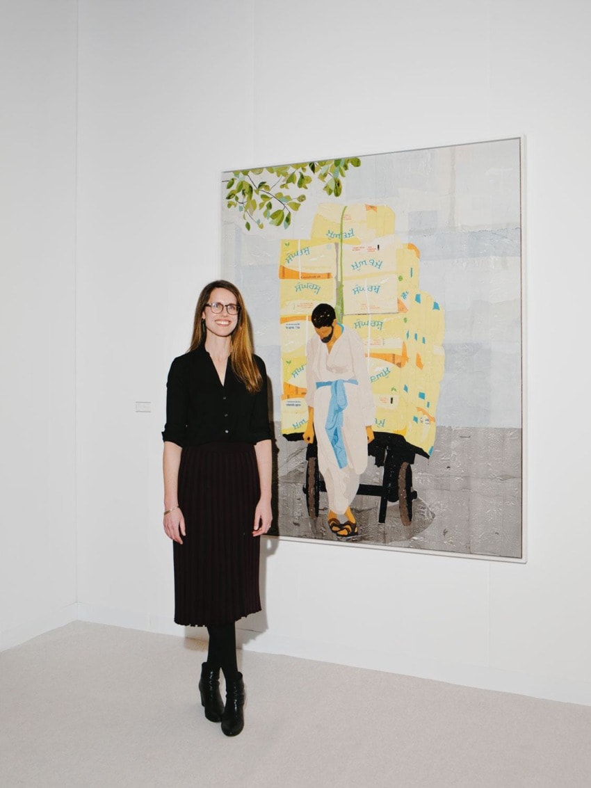 Person standing in art gallery beside a painting of a seated figure surrounded by stacked yellow boxes with patterns.