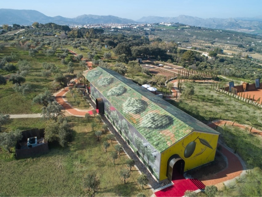 Aerial view of a modern building with a green patterned roof surrounded by trees and pathways in a scenic landscape.