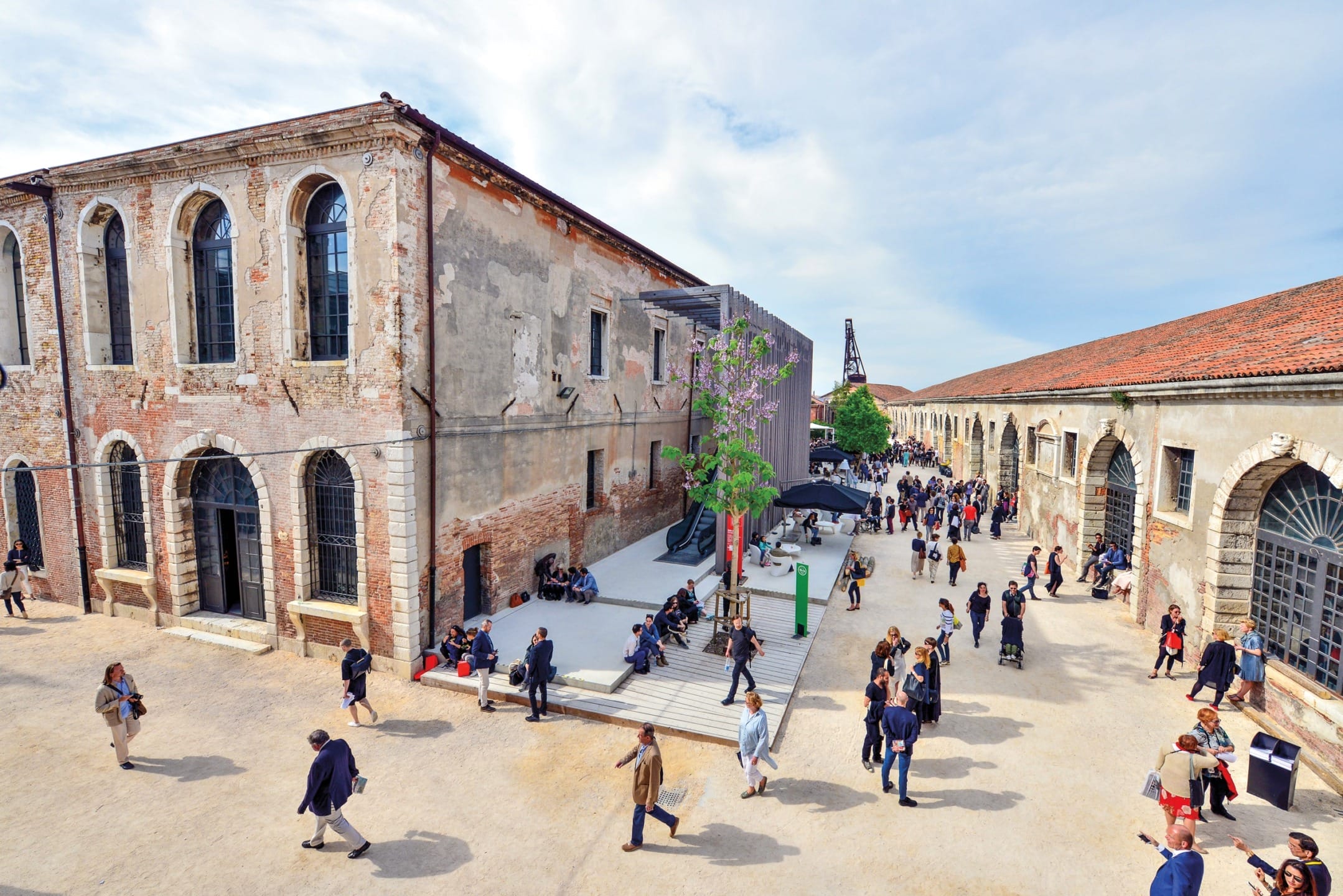 People walking and gathering in an outdoor courtyard with historic brick buildings under a partly cloudy sky.