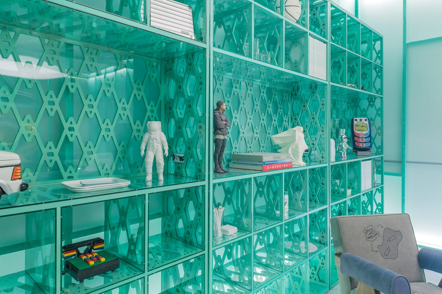 Teal display shelves with various figurines, books, and colorful objects, next to a gray armchair with printed designs.