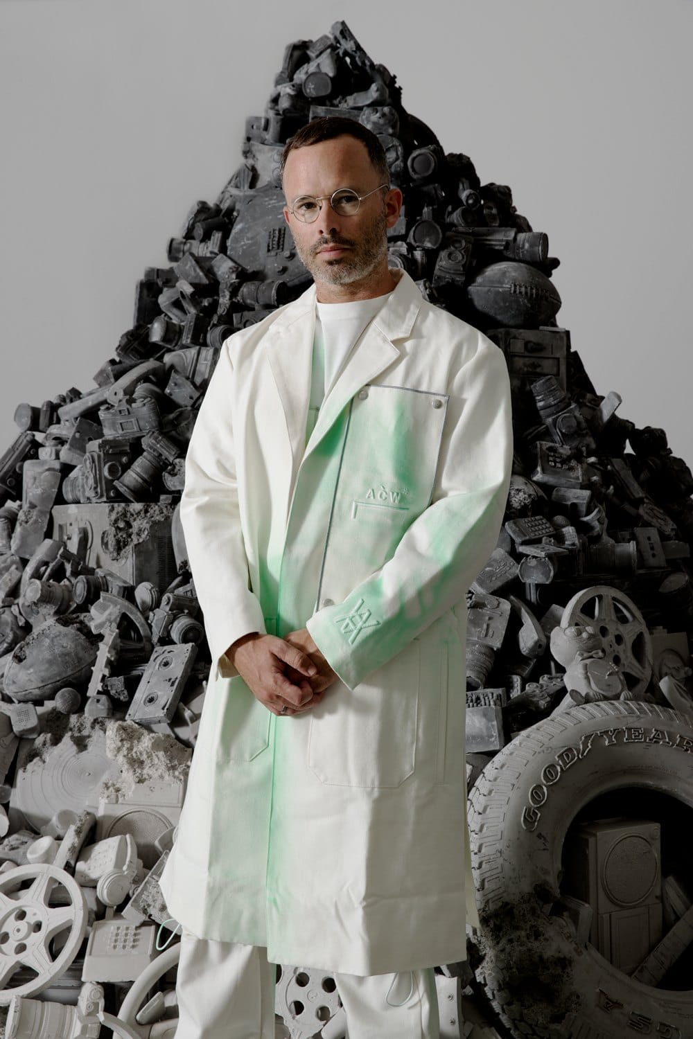 Man in white lab coat with green paint stands in front of a large pile of recycled electronics and mechanical parts.