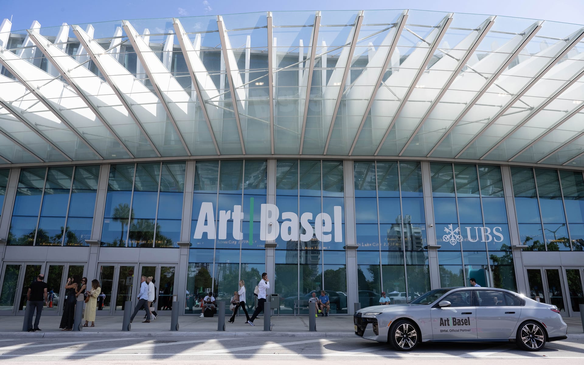 People walking outside the entrance of Art Basel, a car parked in front under a large glass canopy, reflecting clear skies.