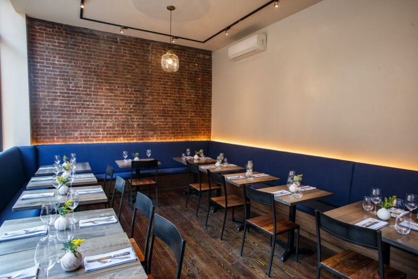 Cozy modern dining room with exposed brick wall, wooden tables set with glassware, and blue cushioned seating.