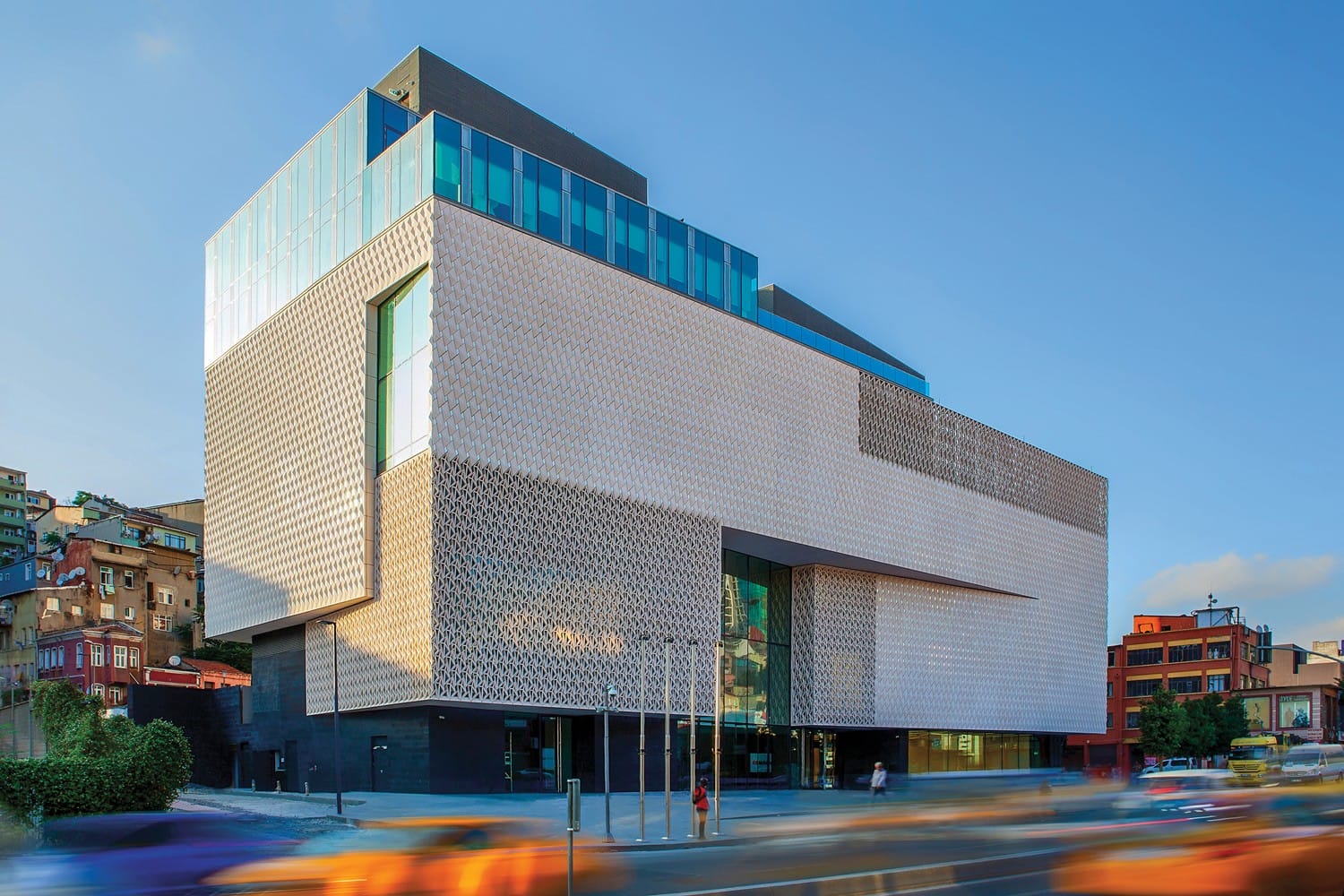 Modern architectural building with textured facade under a clear blue sky, surrounded by blurred motion of passing vehicles.