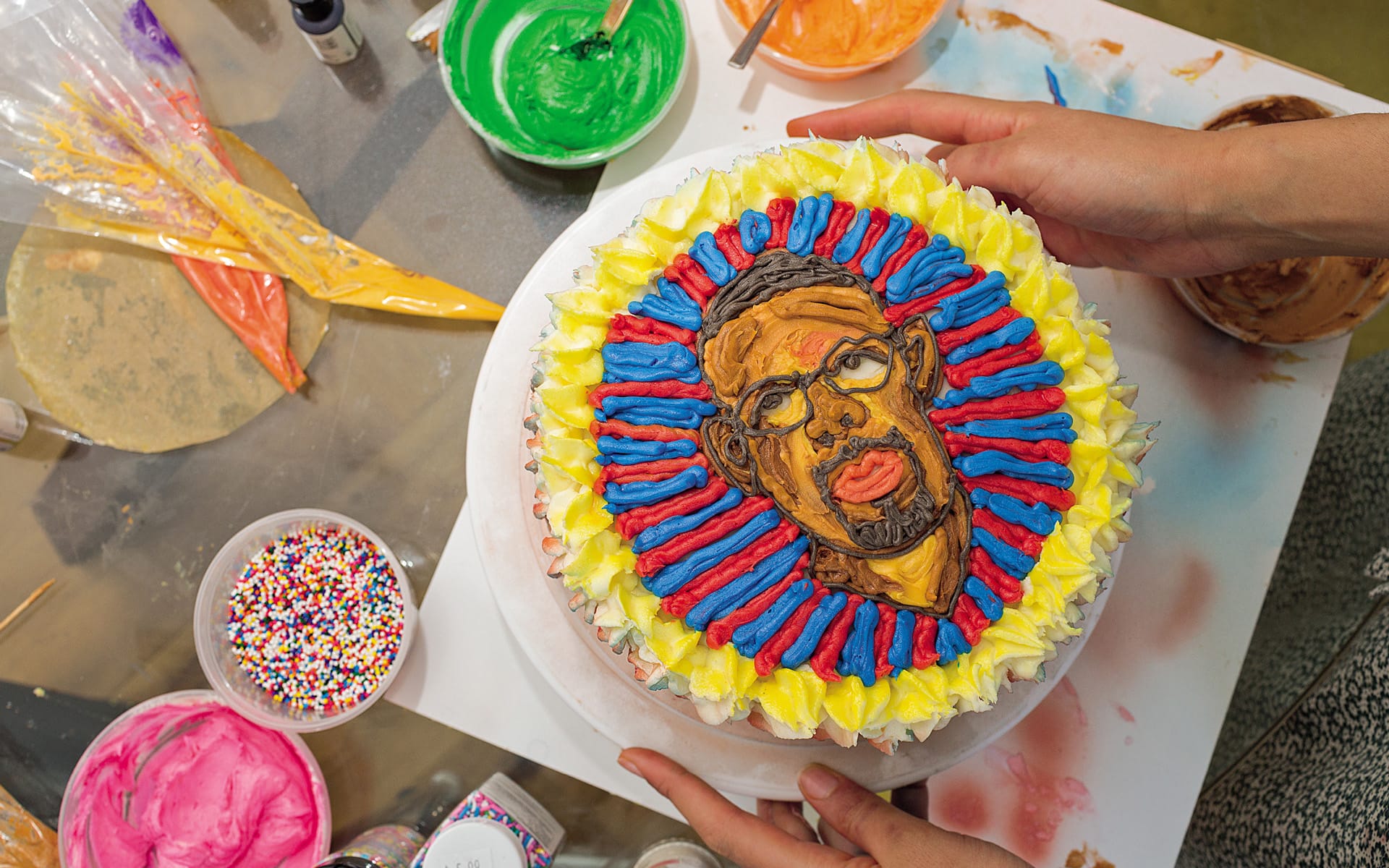 Hands holding a decorated cake with a colorful portrait surrounded by icing and sprinkles on a cluttered table.