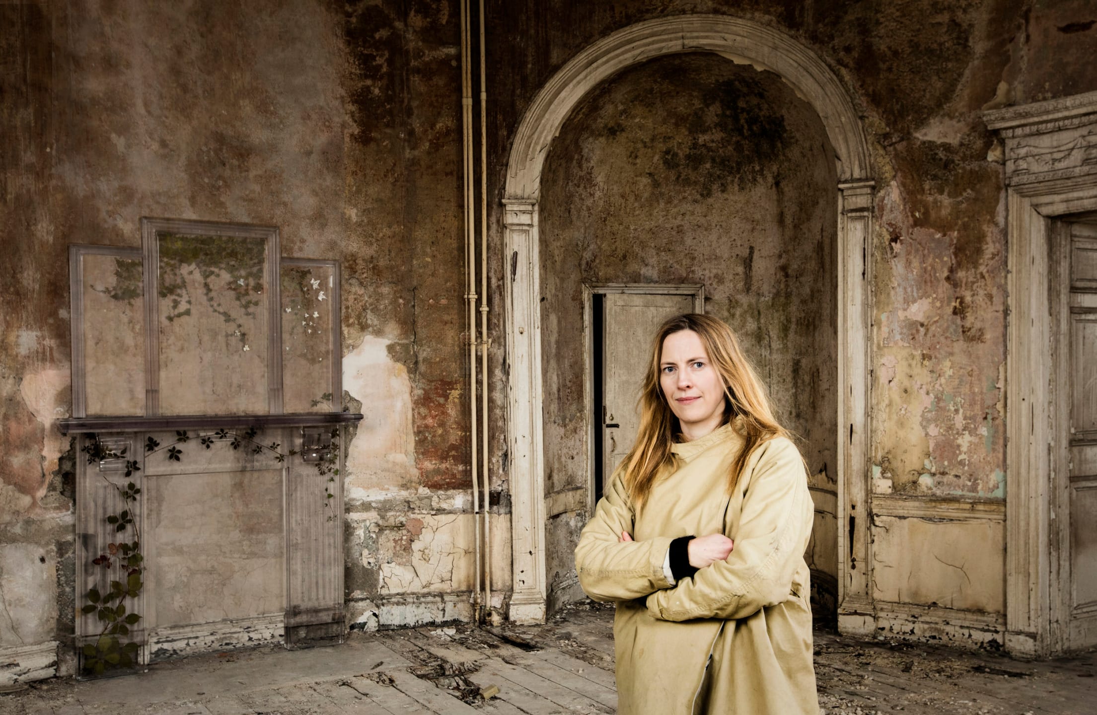 Person in a beige coat standing in an old, weathered room with peeling paint and arched entrance.