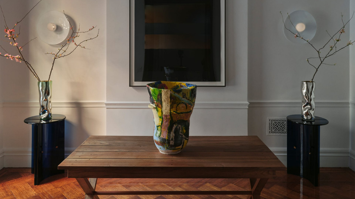 Wooden table with colorful vase, flanked by two sculptural vases on stands with branches, modern art piece on wall behind.