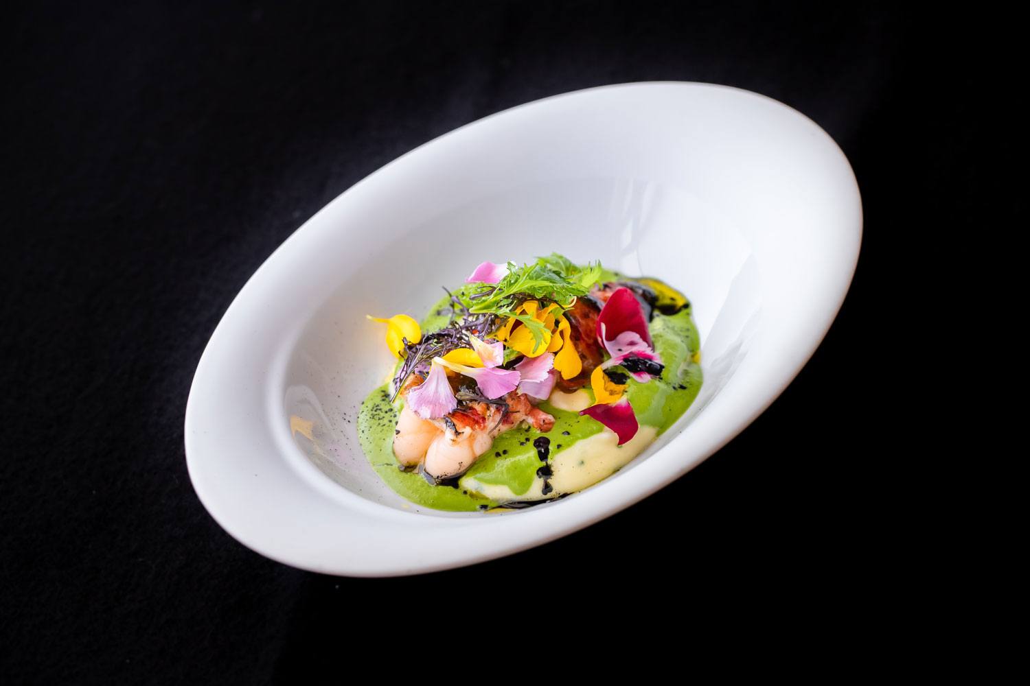 Elegant gourmet dish with colorful flowers, herbs, and sauce on a white plate, presented on a dark background.