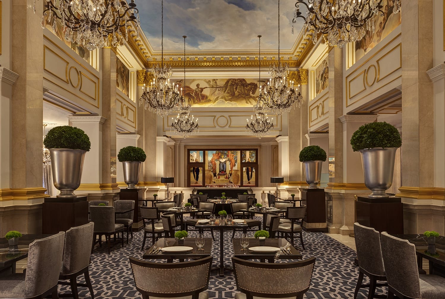 Elegant hotel dining area with ornate chandeliers, classical ceiling art, and neatly arranged tables and chairs.