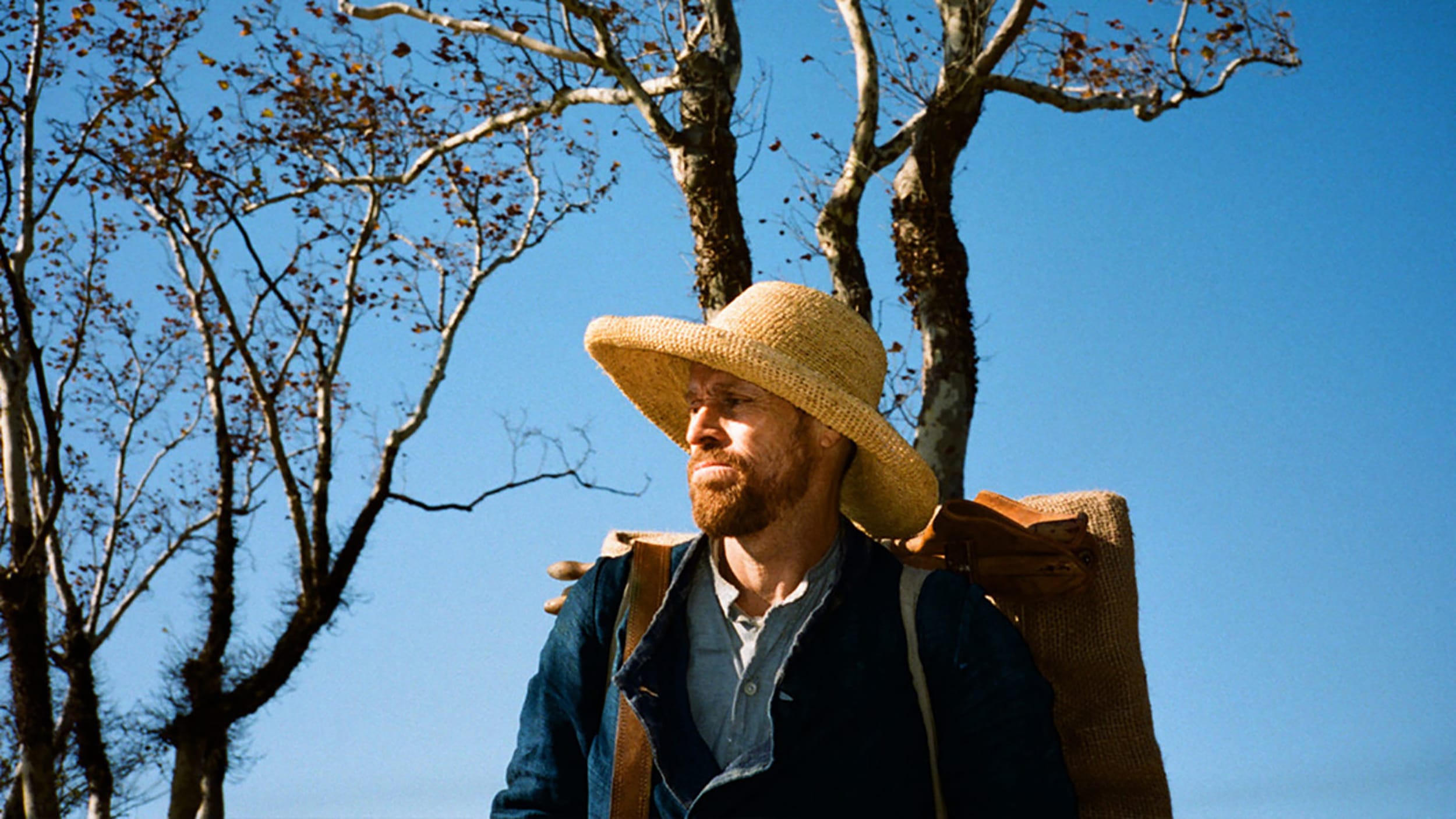 Man wearing straw hat and backpack stands under leafless trees against a clear blue sky.