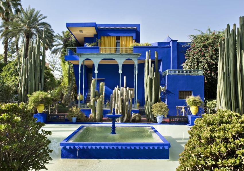 Vibrant blue villa surrounded by lush greenery and tall cacti with a central fountain in a serene garden setting.
