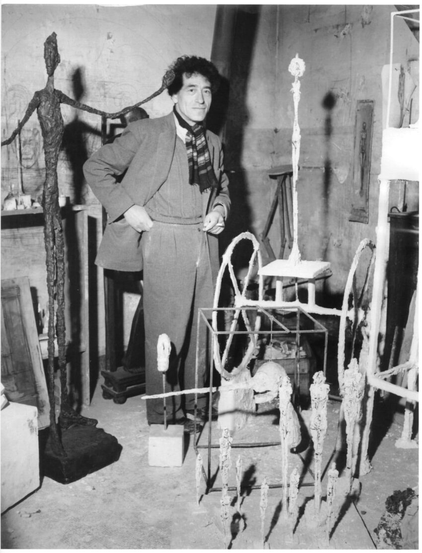 Man in a scarf stands in an art studio surrounded by abstract sculptures.
