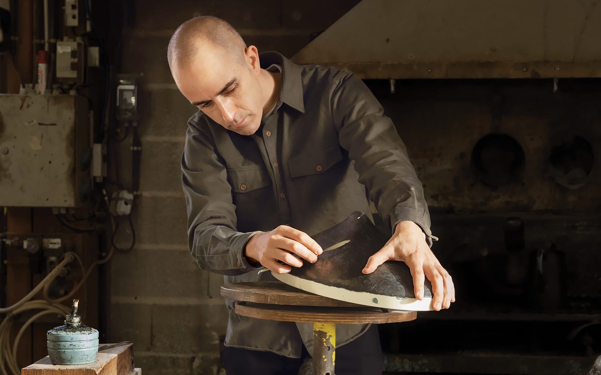 Person crafting a shoe sole in a workshop, focused and using a tool, surrounded by workshop equipment.