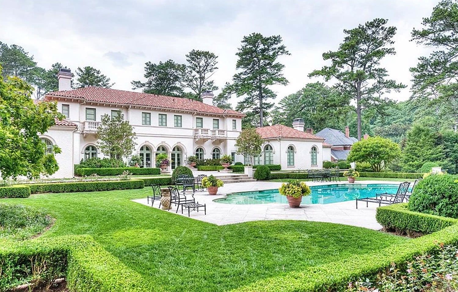 Elegant mansion with red-tiled roof, lush garden, and a pool surrounded by trees and neatly trimmed hedges in the backyard.