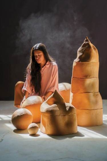 Woman in pink shirt sitting beside large abstract clay sculptures in a sunlit room with a dark background.