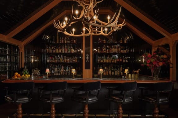 A dimly lit rustic bar with an antler chandelier, dark wood paneling, and a fully stocked back bar.