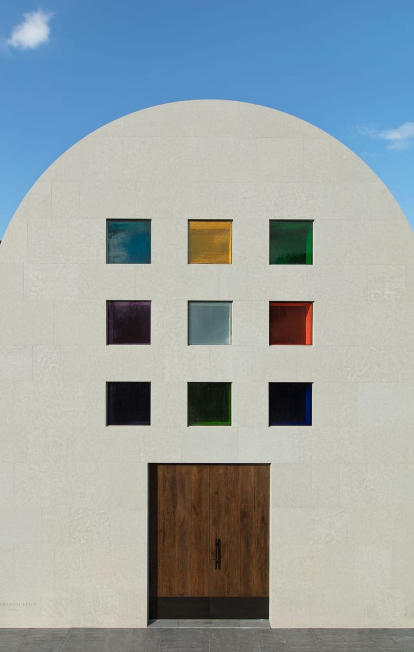 Modern white building facade with nine colorful square windows and a large wooden door under a clear blue sky.