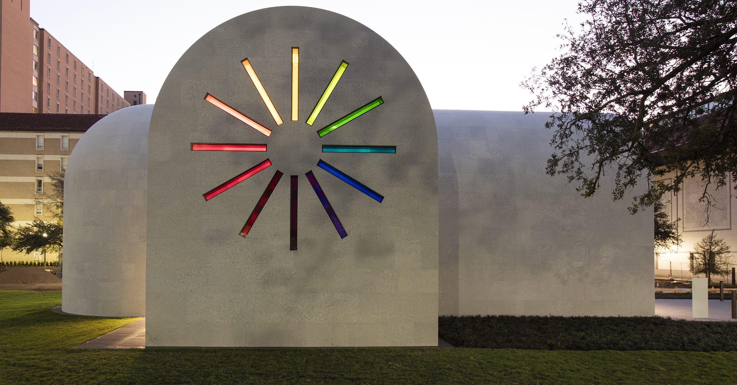 Building facade with colorful stained glass window in starburst pattern surrounded by trees and grass at dusk.