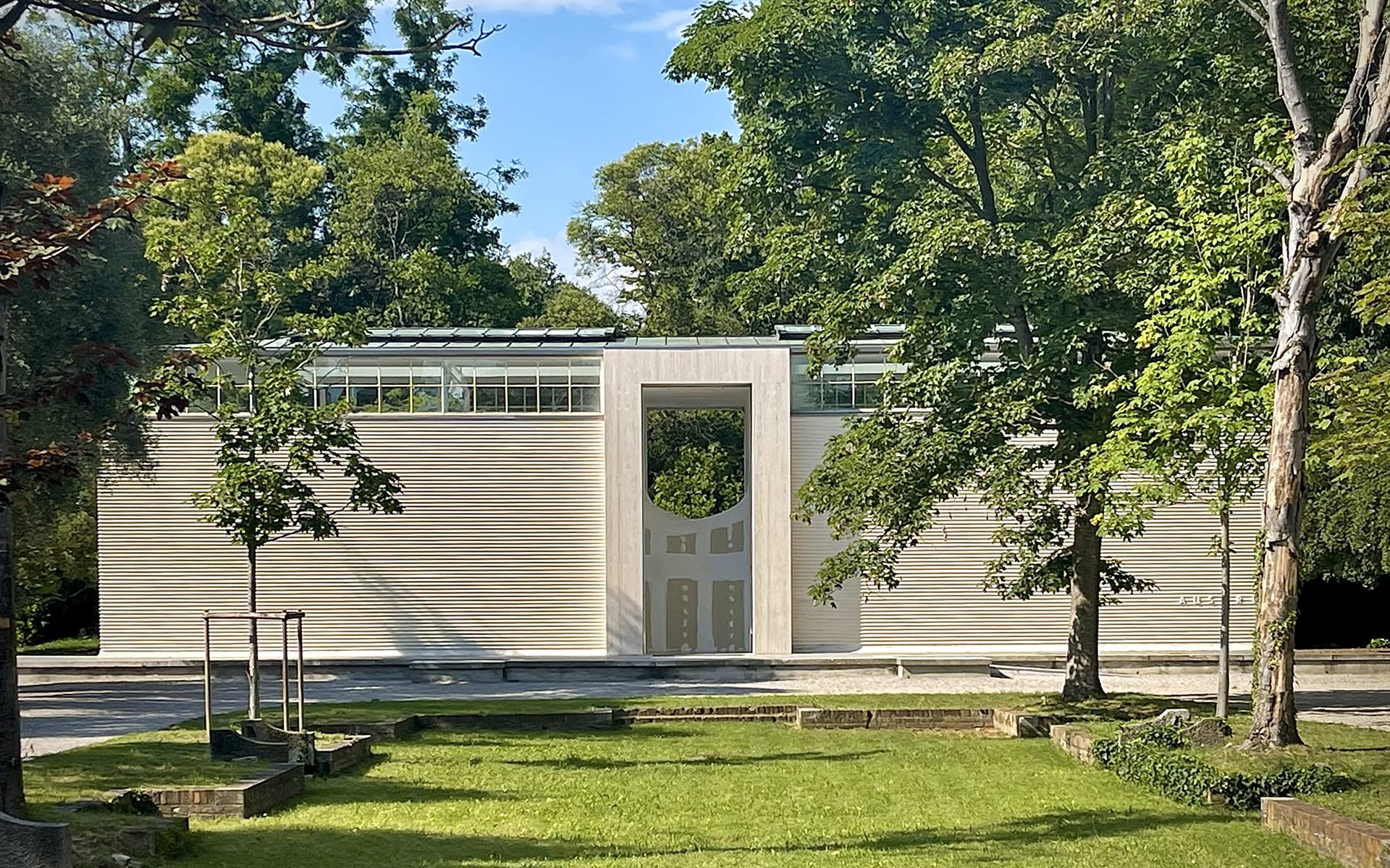 Modern building with a unique archway, surrounded by lush trees and greenery under a clear blue sky.