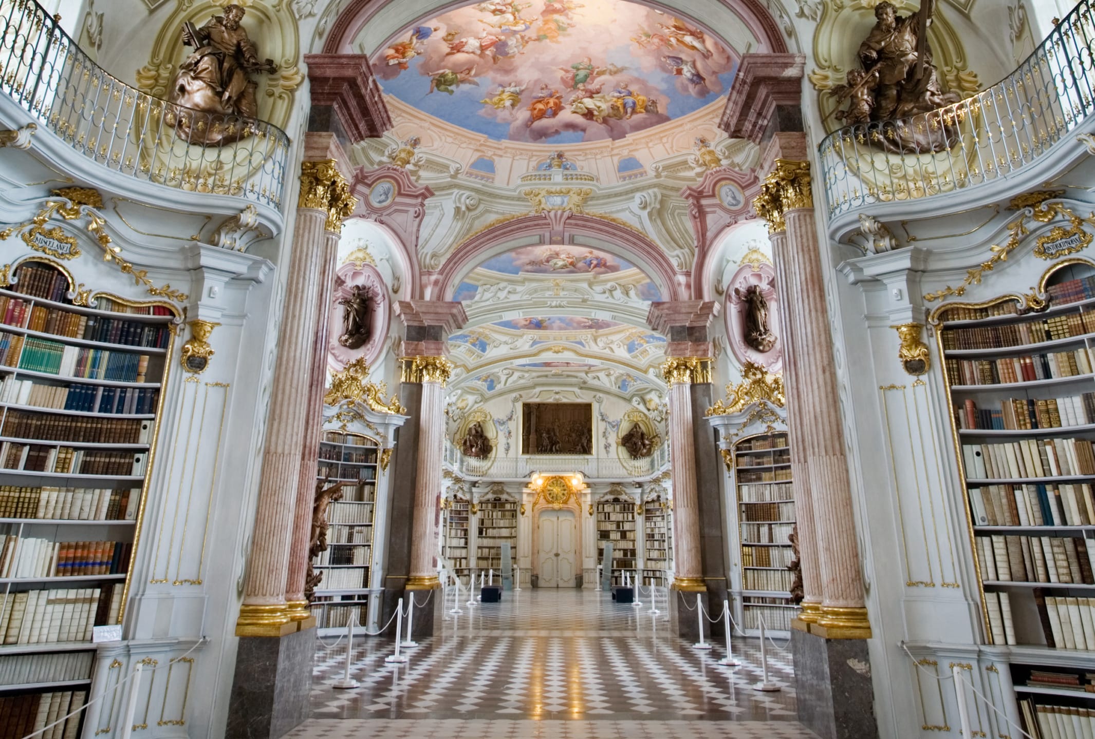 Baroque library interior with ornate ceiling, gilded decorations, and tall bookshelves filled with books.