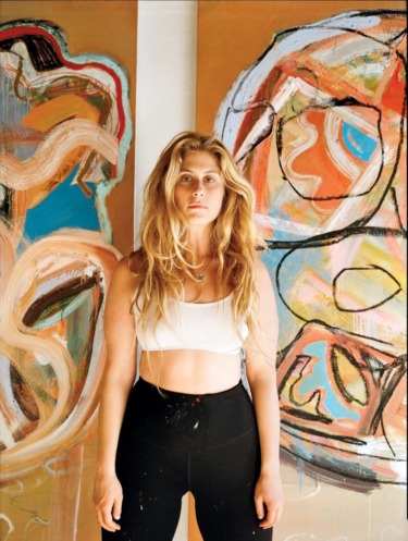 Artist standing in front of abstract paintings with vibrant colors and bold brushstrokes, wearing a white top and black pants.