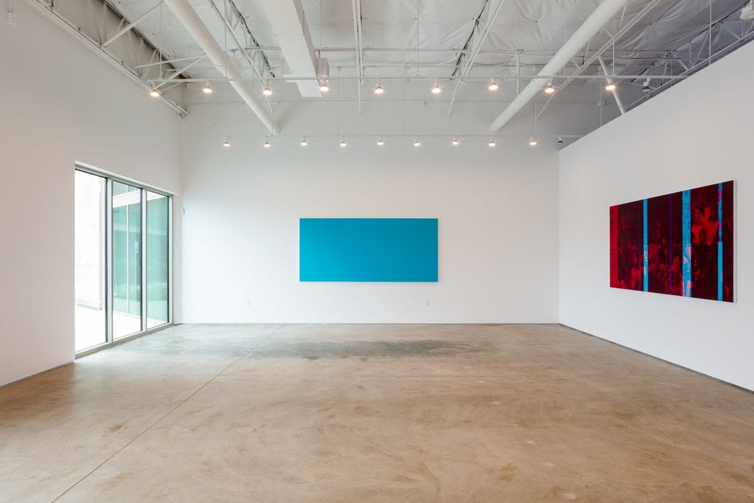 Minimalistic art gallery with two large abstract paintings on white walls, one blue and one red, under bright overhead lighting.