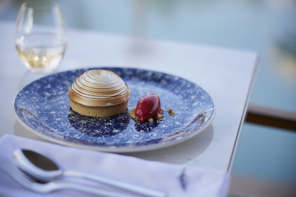 Tart with a swirled meringue top and a scoop of red sorbet on a floral blue plate, set on a white table near a glass of wine.