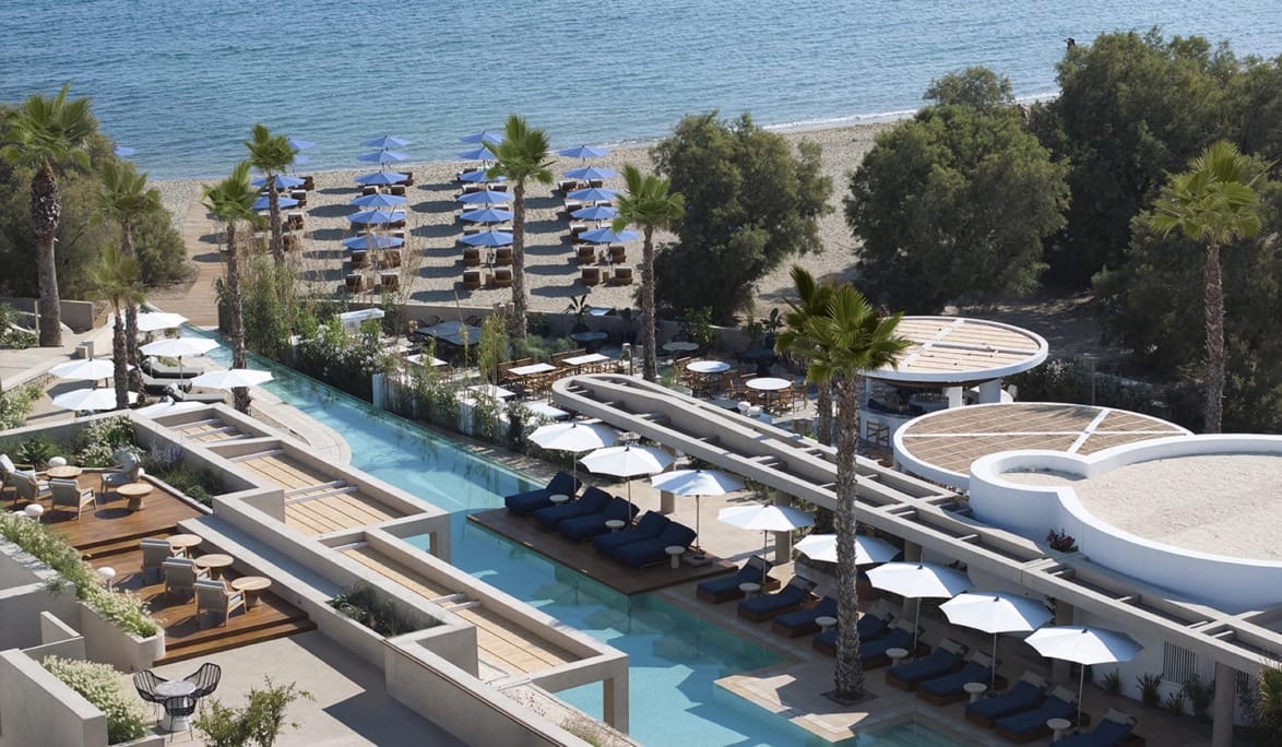 Modern beach resort with poolside lounge chairs, umbrellas, palm trees, and a view of the sea lined with umbrellas and sunbeds.