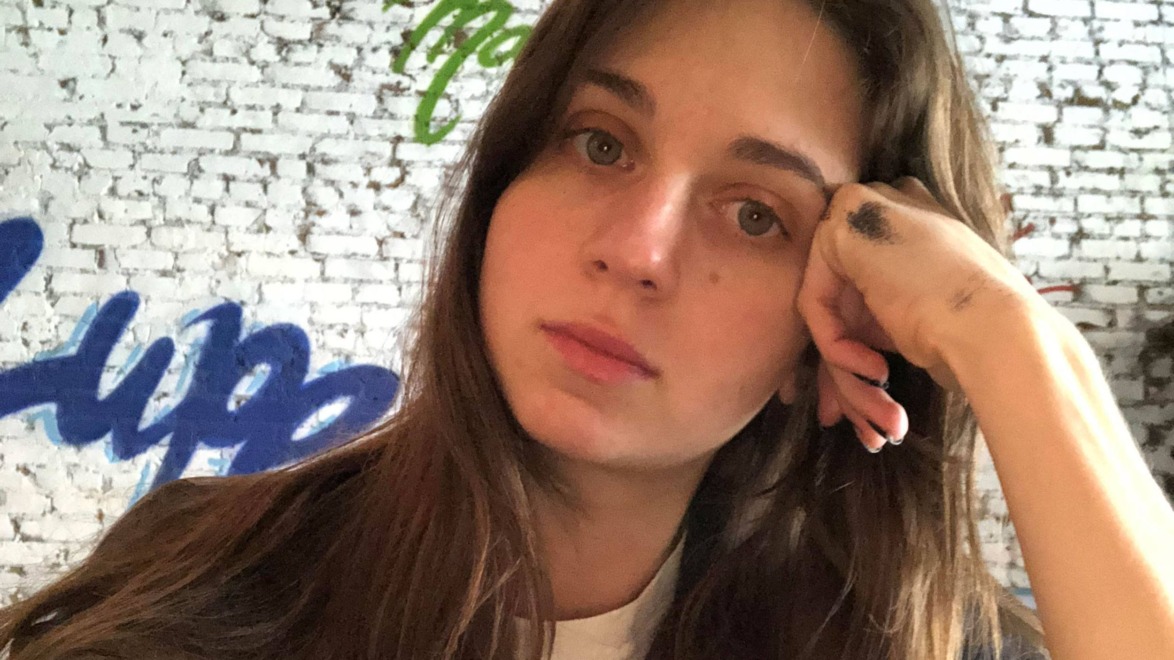 Person sitting indoors, resting their head on their hand, with graffiti on a brick wall in the background.