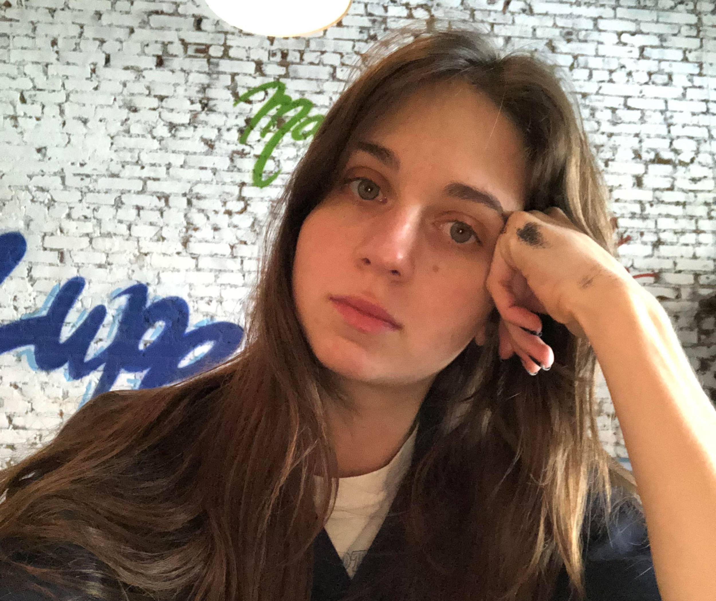 Person sitting indoors, resting their head on their hand, with graffiti on a brick wall in the background.