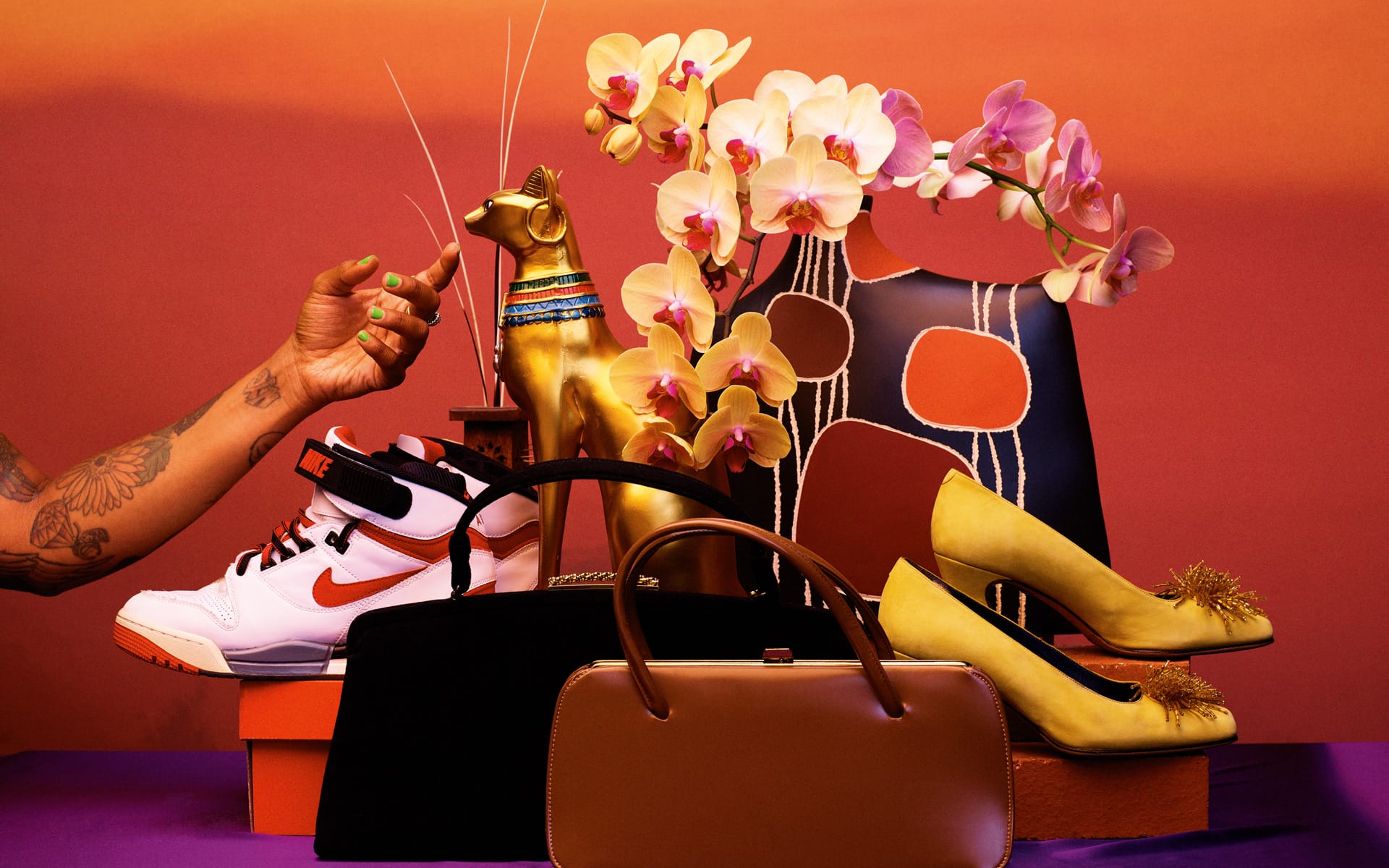 A colorful still life with sneakers, high heels, a handbag, a golden cat statue, and orchids against a vibrant background.