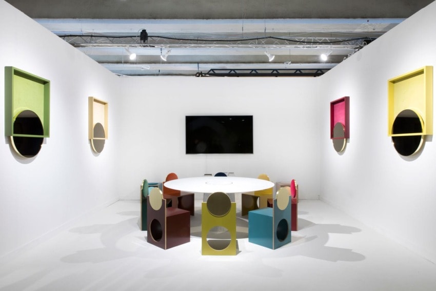 Colorful modern art installation with circular chairs arranged around a table in a white gallery space.